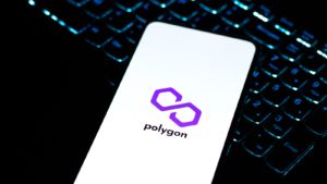 Un teléfono, encima del teclado de una computadora portátil, que muestra el logotipo de Polygon.  Predicciones de precios de polígonos