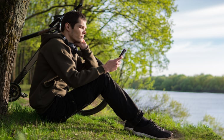 Una persona se sienta en la orilla de un río junto a su bicicleta, mirando su teléfono inteligente.