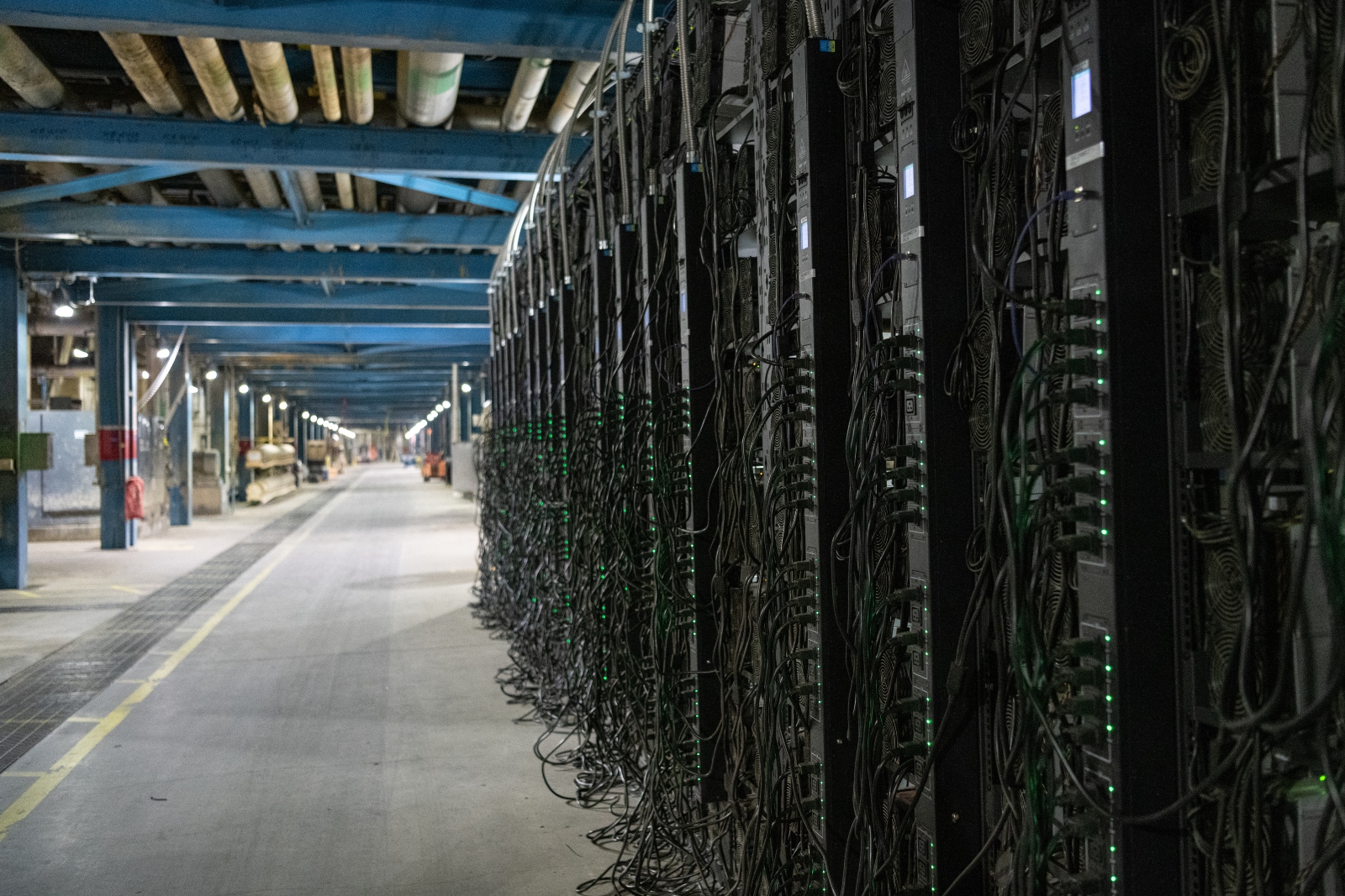 una enorme pared de servidores negros con luces verdes brillantes y cables negros colgando de ellos en un pasillo vacío.