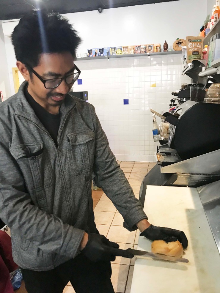 Ryan Natividad viste una chaqueta gris y guantes.  Corta un bagel en la cocina de su tienda, The Bagel Exchange. 