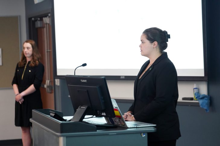 La estudiante de negocios Emma Rolf, a la derecha, habla mientras su compañera de equipo Riley Smith espera para agregar a la presentación del Equipo 1. También estuvieron presentes los miembros del equipo Hope Wilder y Kyle García. (Foto de Suzanne Carr Rossi).