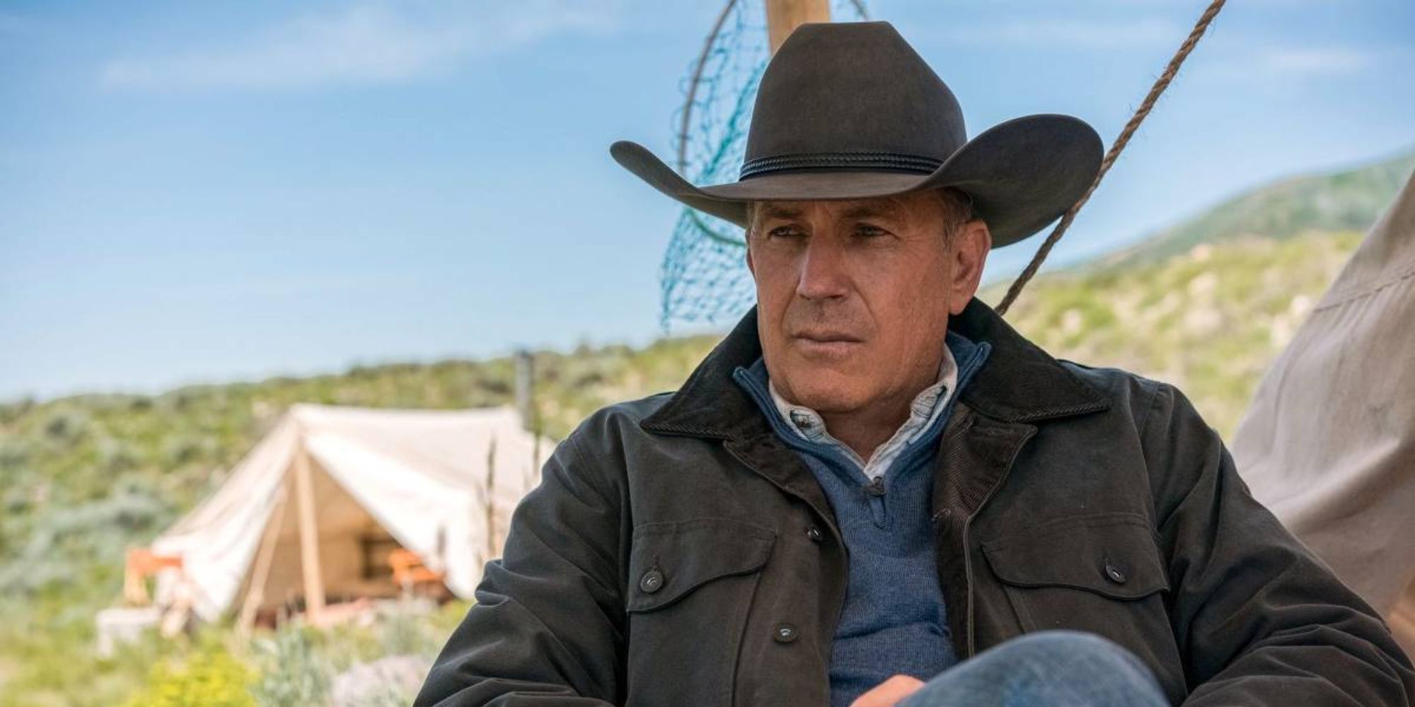 Kevin Costner en Yellowstone sentado con un sombrero de vaquero, una carpa al fondo.