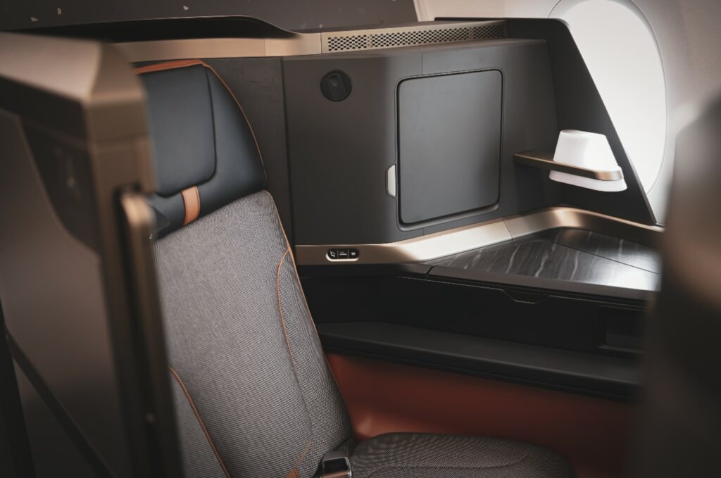 La suite Elements en un asiento Starlux A350.  El asiento es gris, con un reposacabezas negro y acentuado en cobre y naranja quemado.  Un labio como un halo en la cubierta se inclina hacia adentro para mayor privacidad.