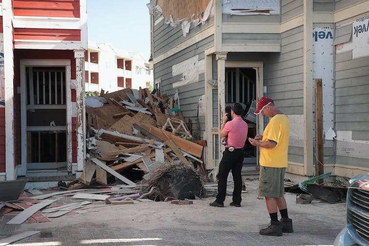 Un hombre con una cinta métrica en el cinturón y una cámara mira los montones de escombros que quedaron entre los edificios después de que el huracán Michael azotara Florida.  El revestimiento del edificio también está arrancado.