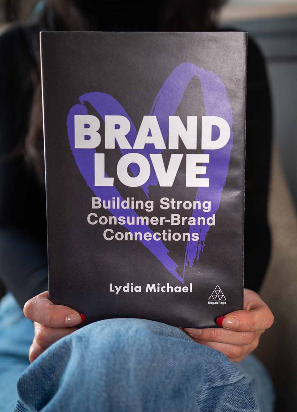 Lydia Michael, ex alumna de Wayne State Ilitch School, sentada en una silla con su primera novela, Brand Love.