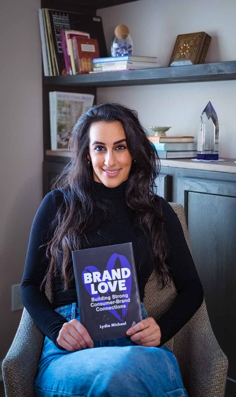 Lydia Michael, ex alumna de Wayne State Ilitch School, sentada en una silla con su primera novela, Brand Love.