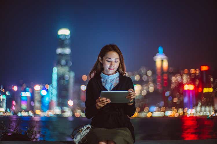 Joven mujer asiática alegre que usa una tableta digital en el paseo marítimo contra el paisaje urbano iluminado por la noche