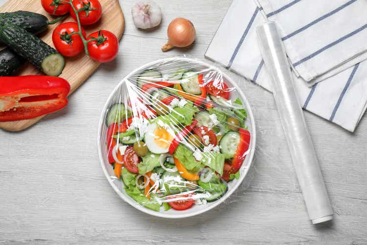 Tazón de ensalada fresca con envoltura de plástico para alimentos sobre una mesa de madera blanca, puesta plana