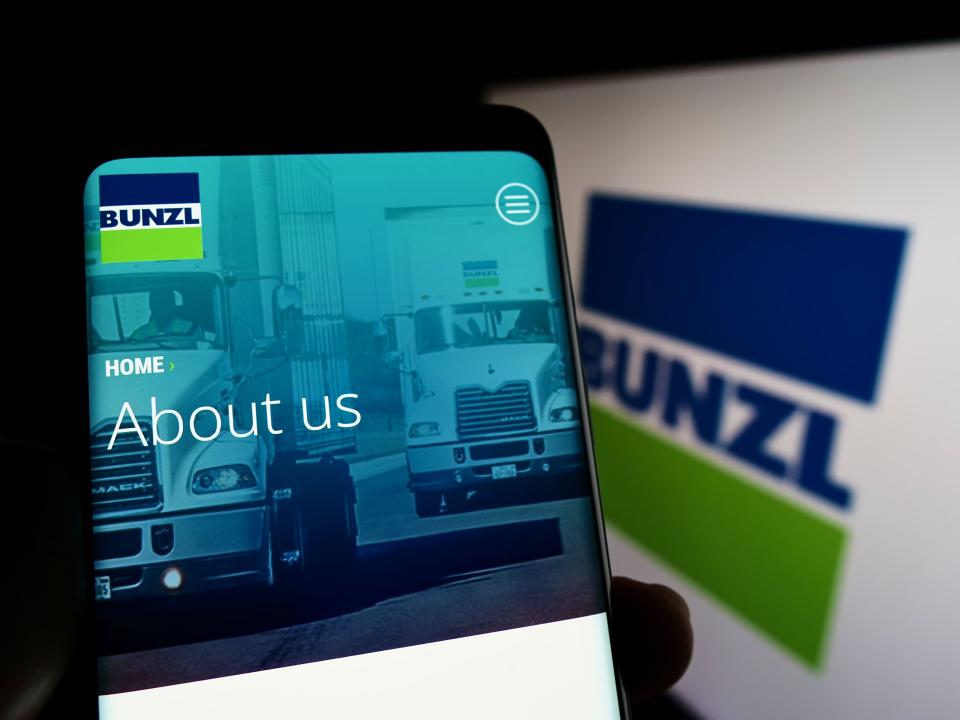 Persona que sostiene un teléfono inteligente con el sitio web de la empresa de distribución británica Bunzl plc en la pantalla frente al logotipo.  Centrarse en el centro de la pantalla del teléfono.