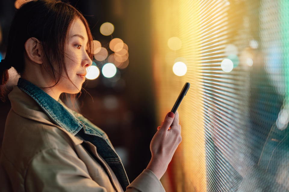 Joven asiática usando un teléfono inteligente contra una pantalla digital iluminada en la calle de la ciudad por la noche.  Tecnología de cadena de bloques.  Comercio de criptomonedas.  Inversión NFT (Token no fungible).