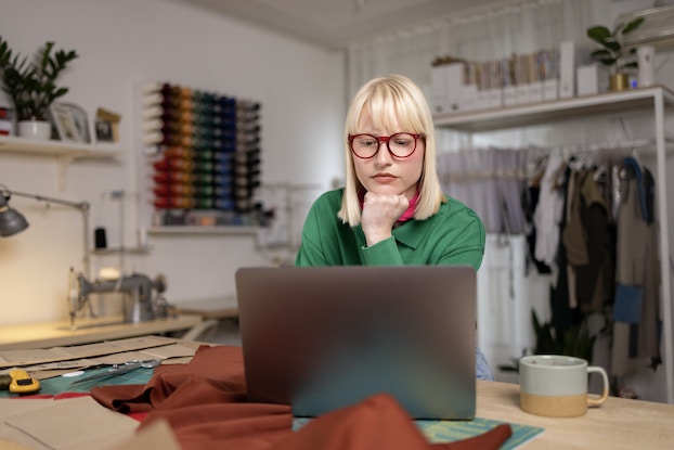     Una mujer sentada en una mesa frente a una computadora portátil abierta.  Mira la pantalla del portátil con la barbilla apoyada en el puño.  Al fondo hay una habitación de paredes blancas con un perchero con ropa a lo largo de una pared y filas de bobinas de hilo de todos los colores montadas en la otra pared.  La mujer tiene el pelo rubio hasta los hombros y viste una camisa verde y gafas con montura roja.