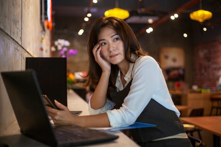 La propietaria de un café sentada frente a su teléfono y su computadora portátil y luciendo preocupada.