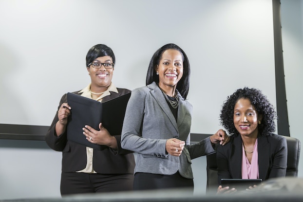     Un trío de mujeres negras vestidas de negocios se enfrenta a la cámara.  Las mujeres están sonriendo.  La mujer de pie a la izquierda sostiene una carpeta abierta y la que está sentada a la derecha sostiene una tableta electrónica.