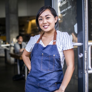 Una mujer joven se encuentra en la entrada de un restaurante o cafetería, apoyada contra la puerta de cristal.  Lleva una camisa a rayas blancas y celestes debajo de un delantal vaquero.  Tiene una mano en la cadera y sonríe con confianza.