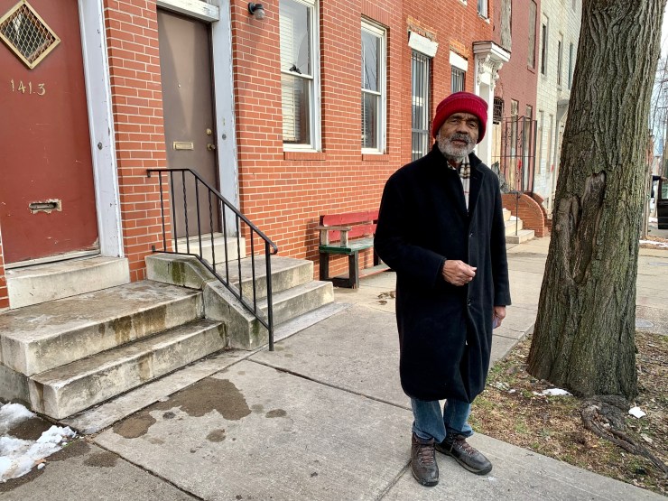 Charles Dugger, un hombre negro que viste un abrigo negro y un sombrero rojo, se encuentra en la acera frente a varias casas en hilera.