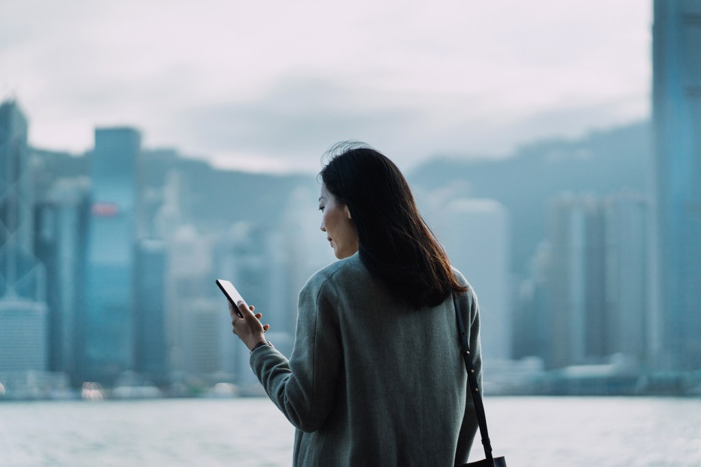 Joven empresaria asiática usando un teléfono inteligente junto al paseo marítimo del puerto Victoria, contra la escena de la ciudad de Hong Kong en el distrito central de negocios