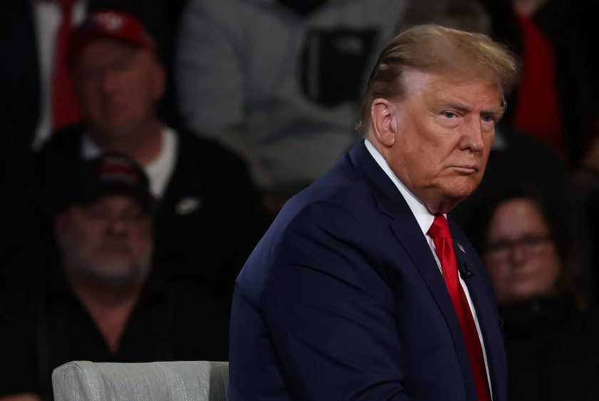 Trump está sentado de lado y tiene una expresión seria.  Lleva un traje oscuro, camisa blanca y corbata roja.