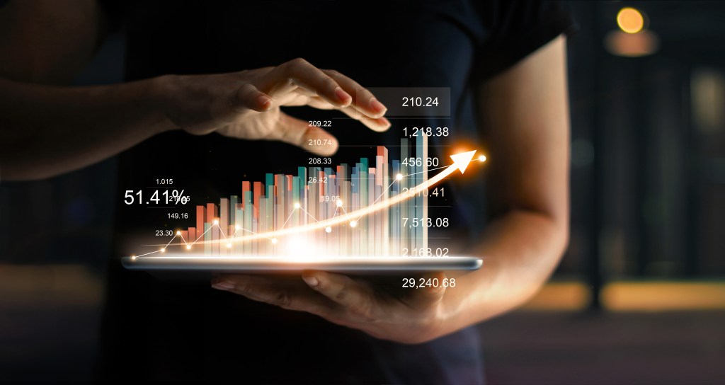 Empresario sosteniendo una tableta y mostrando un creciente holograma virtual de estadísticas, gráficos y tablas con una flecha hacia arriba sobre un fondo oscuro.  Bolsa de Valores.  Concepto de estrategia, planificación y crecimiento empresarial.