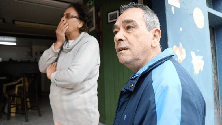 João Batista Coelho, con una chaqueta deportiva azul, está afuera de su tienda, mientras su esposa mira, con una mano sobre la boca, como abrumada por la emoción.