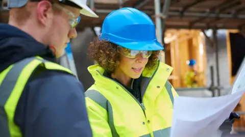 Getty Images Un hombre y una mujer con cascos y ropa de alta visibilidad mirando planos de construcción en una obra.