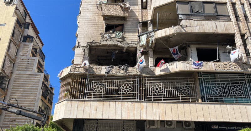 Un edificio de varios pisos dañado con una fachada parcialmente derrumbada. Se ven escombros en el balcón y varias banderas y prendas de vestir cuelgan de la barandilla del balcón. El cielo está despejado y azul al fondo.