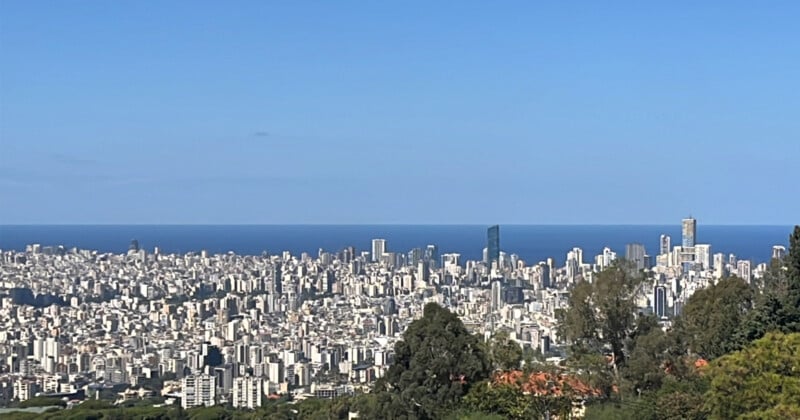 Una vista panorámica de un paisaje urbano con numerosos edificios altos bajo un cielo azul claro. La ciudad se extiende hacia un mar inmenso y tranquilo al fondo. En primer plano se ve una exuberante vegetación.