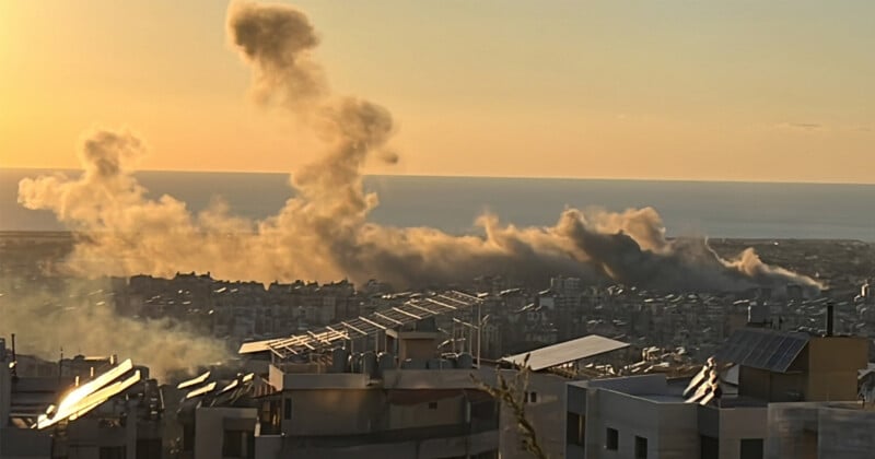 El humo se eleva desde los edificios en un paisaje urbano, iluminado por el sol poniente. En primer plano se ven los tejados con paneles solares y el mar se extiende en el horizonte.