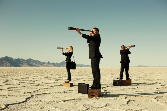 Tres personas vestidas de negocios, de pie sobre cajas y mirando a través de telescopios en diferentes direcciones en el desierto. 