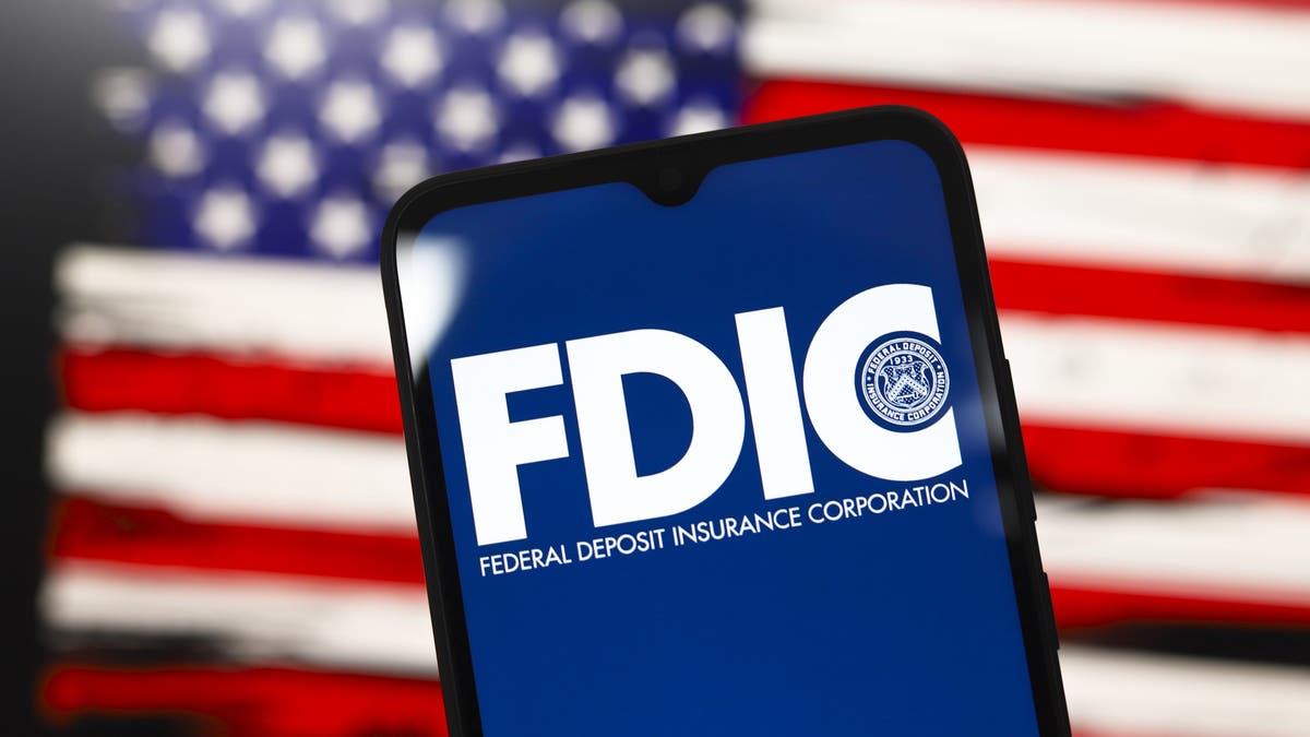 El logotipo de la Corporación Federal de Seguros de Depósitos de los Estados Unidos aparece en la pantalla de un teléfono inteligente y, al fondo, la bandera de los Estados Unidos está en la pantalla de la computadora en Reno, Nevada, el 3 de diciembre de 2024.