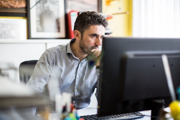     Un hombre está sentado en un escritorio frente a una computadora de escritorio. Tiene una expresión seria en su rostro. Está mirando datos en su computadora y pensando.